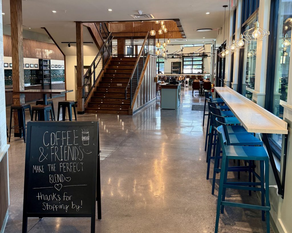 A modern coffee shop interior with high tables and chairs, a chalkboard sign in front, and a wooden staircase leading to an upper level in the background.