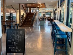 A modern coffee shop interior with high tables and chairs, a chalkboard sign in front, and a wooden staircase leading to an upper level in the background.