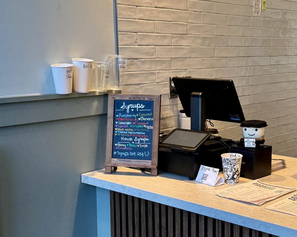 A coffee shop counter with a chalkboard sign listing syrup flavors, several cups on the counter, a cash register, and a tip jar.