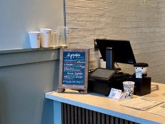A coffee shop counter with a chalkboard sign listing syrup flavors, several cups on the counter, a cash register, and a tip jar.