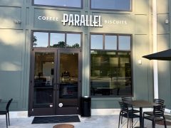 The exterior of Parallel, a café offering coffee and biscuits, with glass doors, windows, and outdoor seating with black chairs and tables.