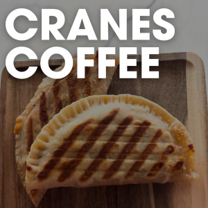Two grilled empanadas on a wooden board tempt your taste buds beneath the bold white letters “Cranes Coffee” at the top—a breakfast worth waking up for in Acworth.