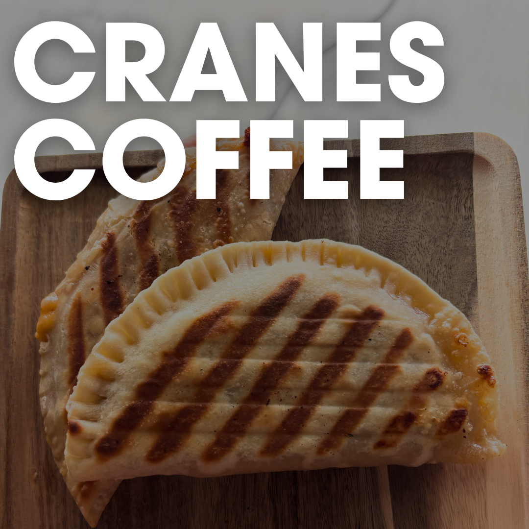 Two grilled empanadas on a wooden board tempt your taste buds beneath the bold white letters “Cranes Coffee” at the top—a breakfast worth waking up for in Acworth.