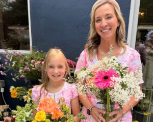 An adult and a child, both in pink dresses, stand outside holding colorful flower arrangements and smiling at the camera.