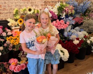Two young girls stand in front of assorted flower bouquets, each holding a small bouquet of orange roses and baby’s breath, smiling at the camera.
