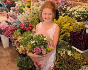 A young girl in a light pink dress holds a bouquet of flowers and smiles, standing in front of shelves filled with various colorful floral arrangements.