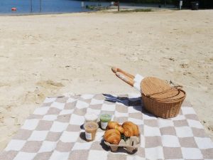 Checkered picnic blanket on sandy beach with a wicker basket, three pastries, and three drinks near a calm body of water—it's a breakfast worth waking up for in Acworth.