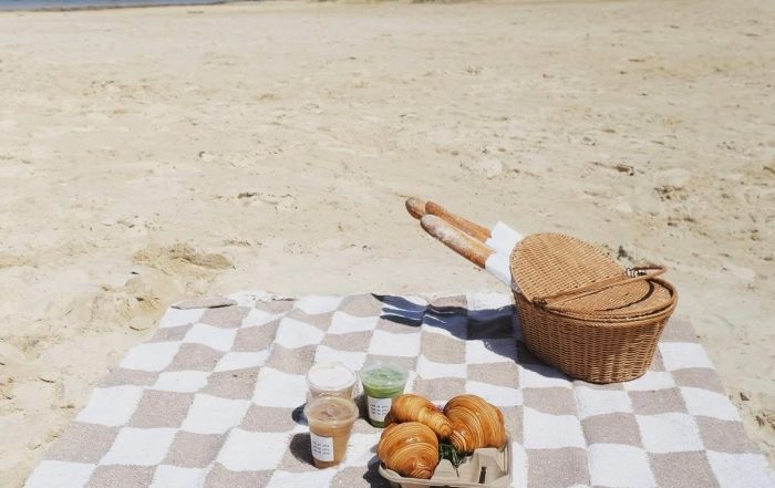Checkered picnic blanket on sandy beach with a wicker basket, three pastries, and three drinks near a calm body of water—it's a breakfast worth waking up for in Acworth.