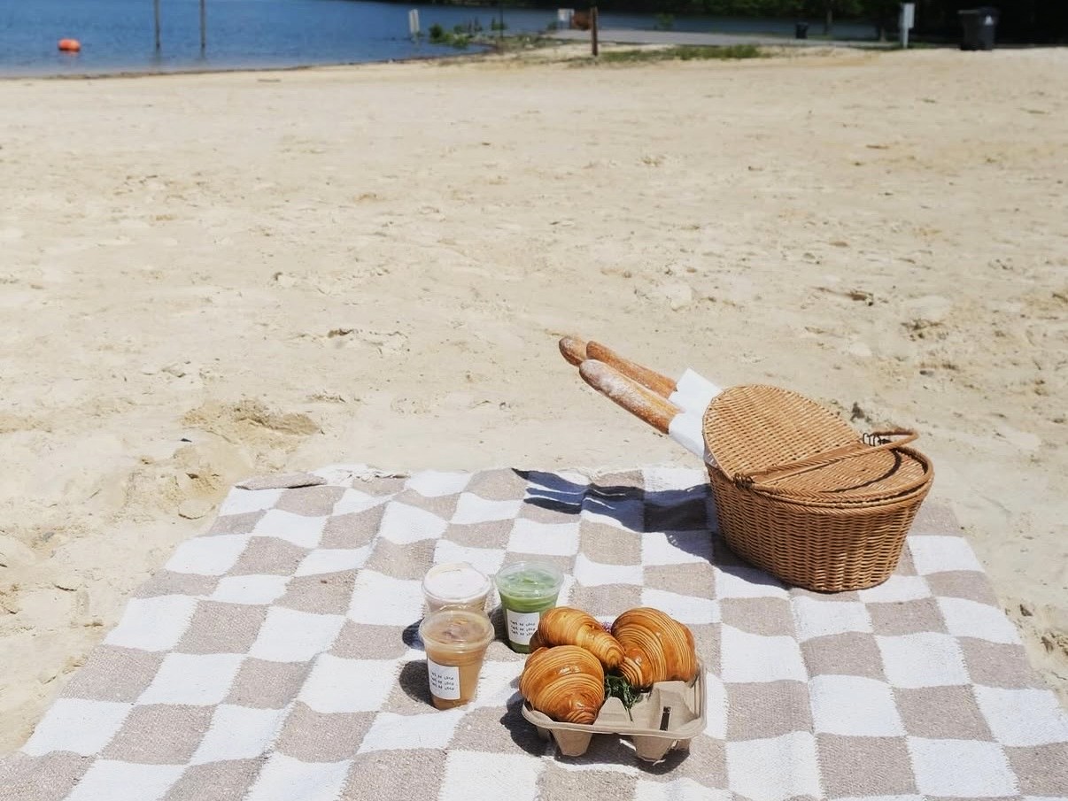 Checkered picnic blanket on sandy beach with a wicker basket, three pastries, and three drinks near a calm body of water—it's a breakfast worth waking up for in Acworth.
