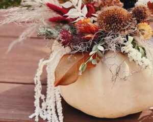 A white pumpkin is used as a vase, holding an arrangement of dried flowers, grasses, and leaves, displayed on a wooden surface.