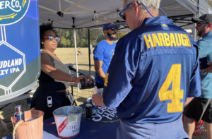 A woman pours a drink for a man wearing a “Harbaugh 4” jersey at an outdoor event booth displaying beer signs.