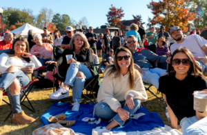 A group of people sit on chairs and blankets outdoors at a festival, smiling and holding drinks, with other attendees and autumn trees in the background.