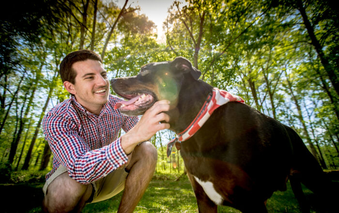 A man in a plaid shirt kneels in a grassy area, smiling and petting a large black dog wearing a red and white bandana, with trees and sunlight in the background—a peaceful moment that feels like breakfast worth waking up for in Acworth.