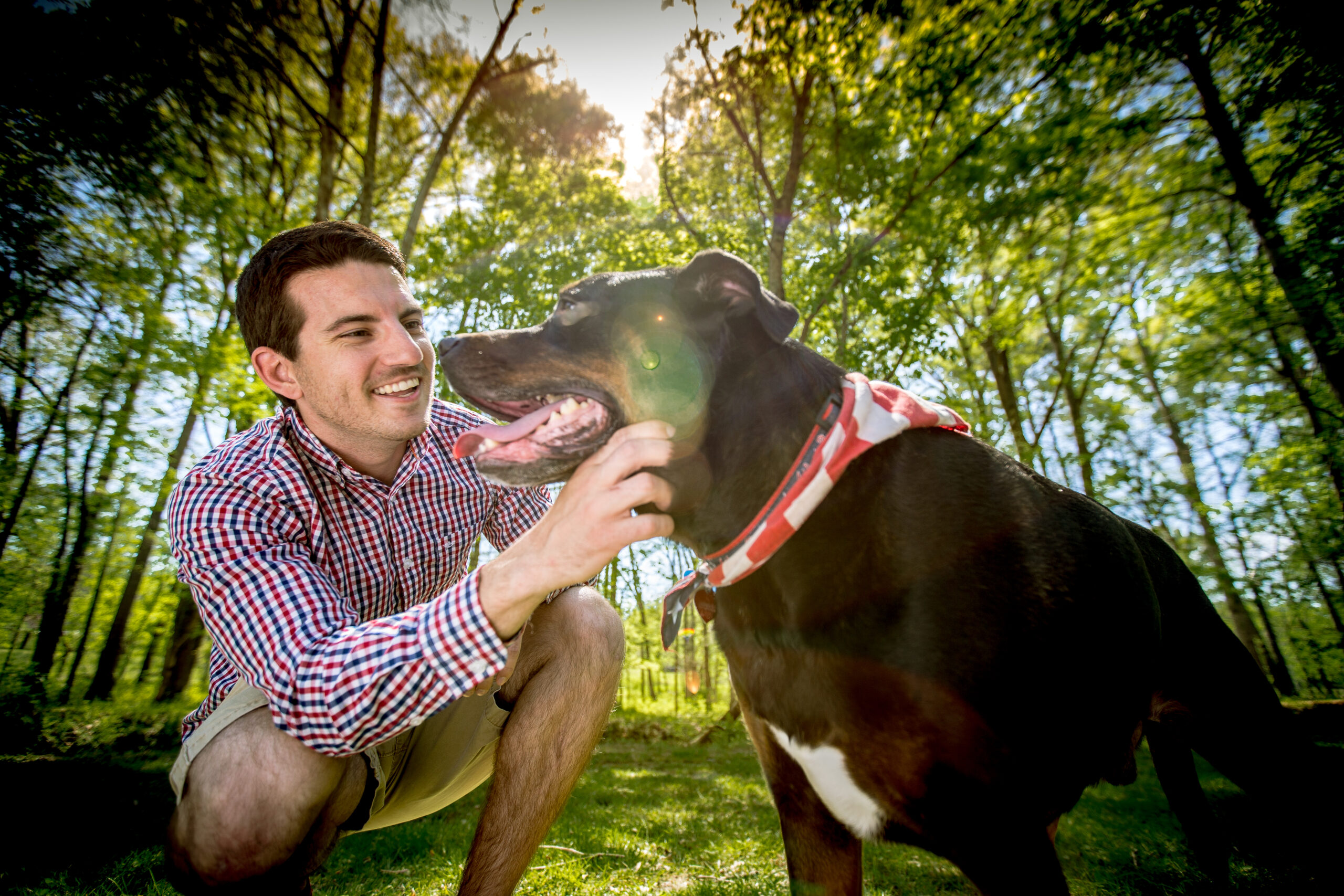 A man in a plaid shirt kneels in a grassy area, smiling and petting a large black dog wearing a red and white bandana, with trees and sunlight in the background—a peaceful moment that feels like breakfast worth waking up for in Acworth.