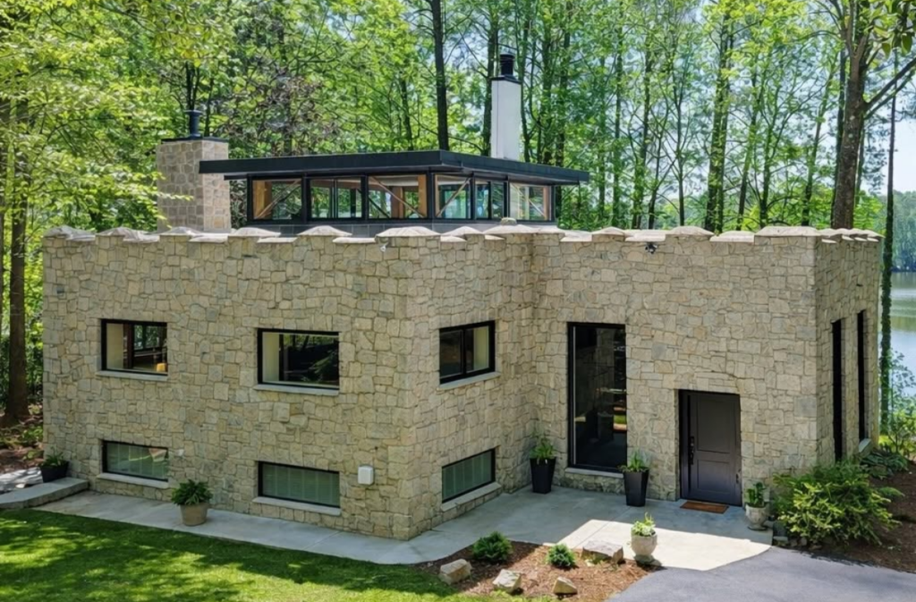 A two-story stone house with a castle-style crenelated roofline and a modern glass clerestory addition, situated in a wooded area near water.