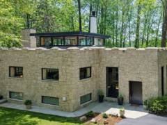 A two-story stone house with a castle-style crenelated roofline and a modern glass clerestory addition, situated in a wooded area near water.