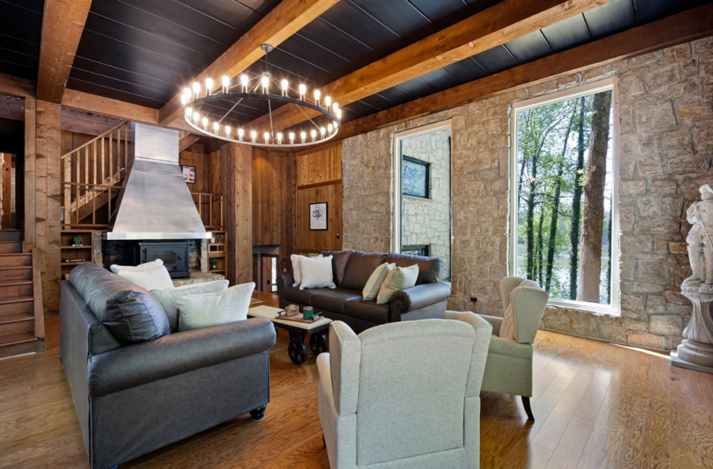 Living room with two leather sofas, two armchairs, a central coffee table, and a large circular chandelier in front of a stone fireplace and windows.