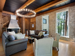 Living room with two leather sofas, two armchairs, a central coffee table, and a large circular chandelier in front of a stone fireplace and windows.