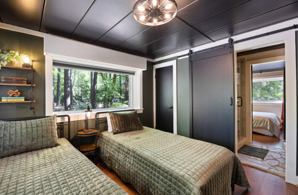 A bedroom with two twin beds featuring quilted olive green bedspreads, a window overlooking trees, and a sliding barn door leading to an ensuite bathroom.
