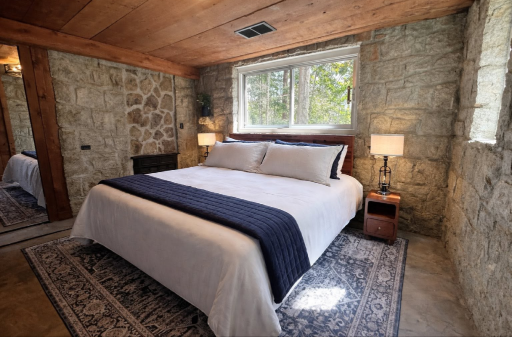 A bedroom with stone walls, a wooden ceiling, and a bed featuring white bedding with a navy blue runner, flanked by two lamps on wooden nightstands.