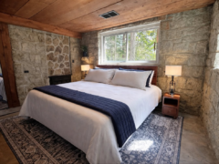 A bedroom with stone walls, a wooden ceiling, and a bed featuring white bedding with a navy blue runner, flanked by two lamps on wooden nightstands.
