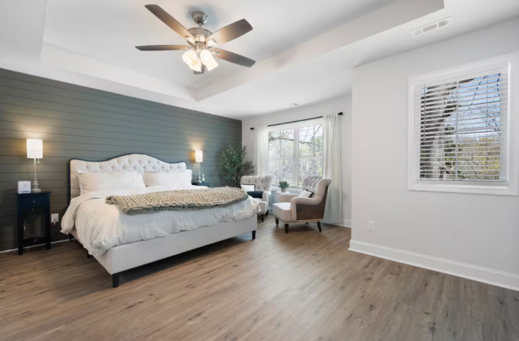 Bedroom with a tufted white bed, an accent wall with gray horizontal paneling, and two armchairs by a window.